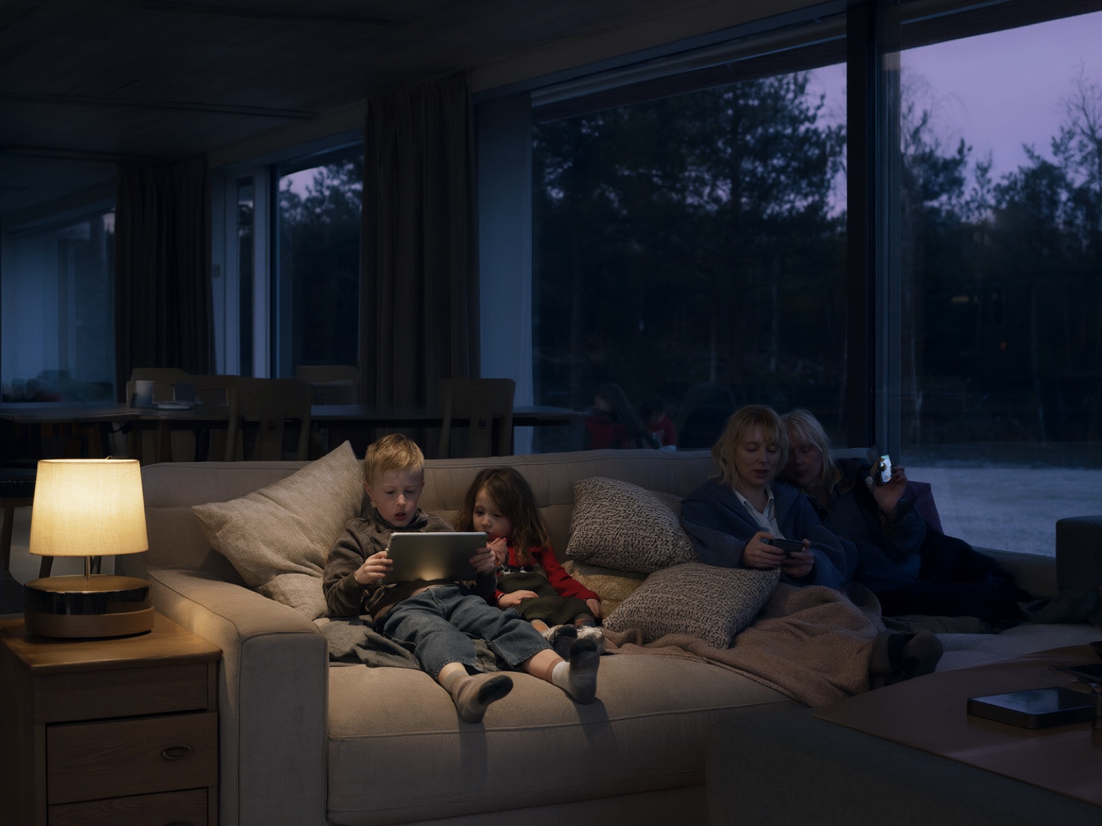 A family relaxing on a sofa in a holiday lodge at dusk, using a tablet and phones
