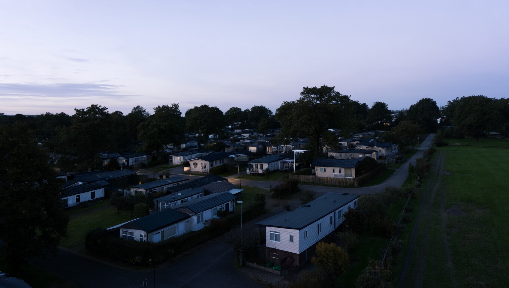 A holiday park at dusk seen from above: rows of static caravans among mature trees, with an open field alongside