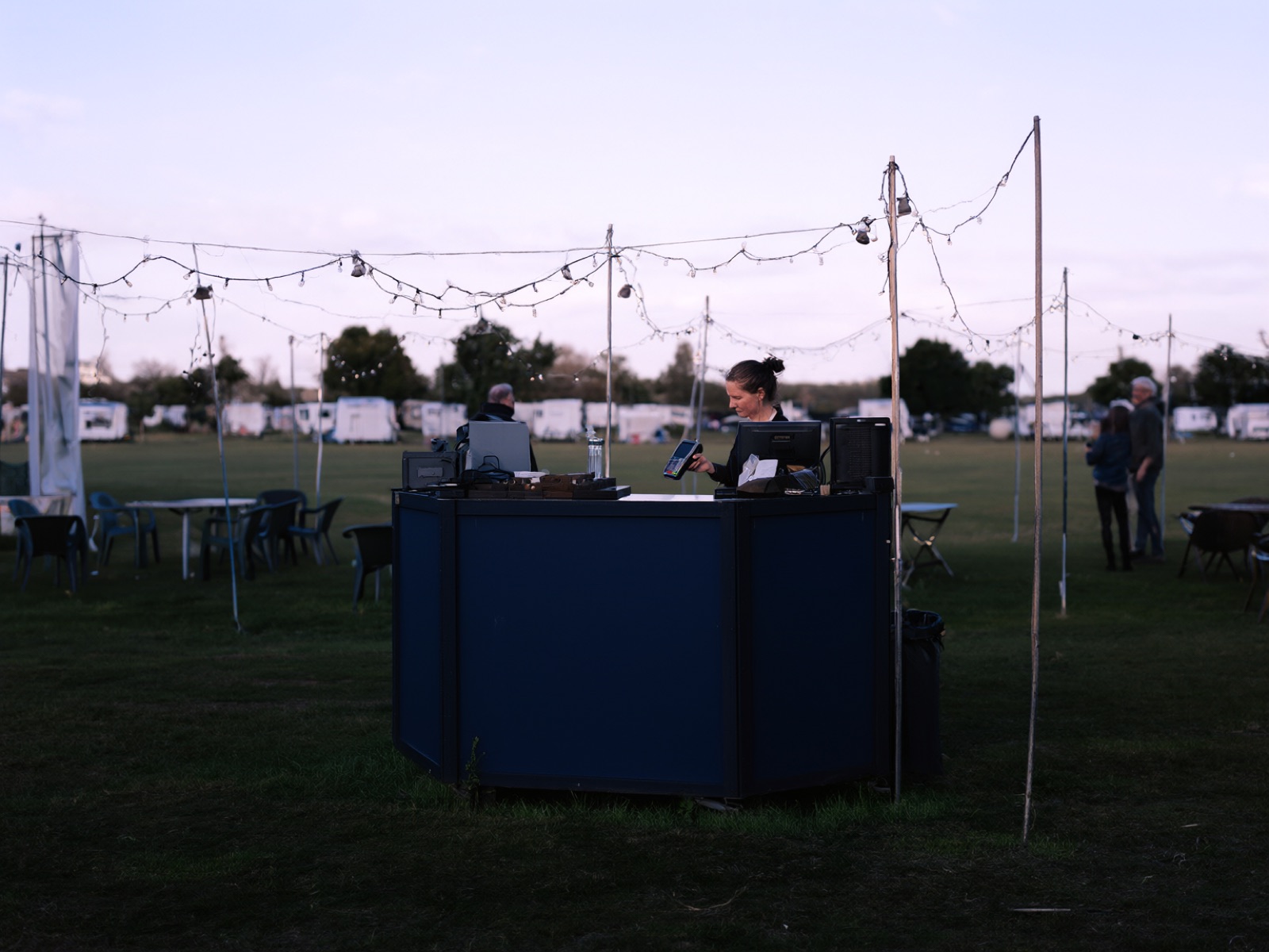 A pop-up bar in a field at dusk under festoon lights, with a staff member holding out a card payment terminal
